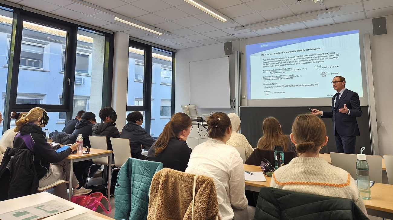 Blick von hinten in einen Hörsaal mit Schüler:innen, die dem Vortrag eines Professors folgen.