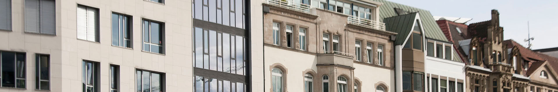 Das Bild zeigt eine Reihe von Gebäuden mit unterschiedlichen architektonischen Stilen. Einige Gebäude haben ein modernes Design mit großen Glasfenstern, während andere älter und aufwendig gestaltet sind – mit detaillierten Fassaden und traditionellen Fenstern. Auch die Dächer unterscheiden sich: Es gibt sowohl Flachdächer als auch geneigte Dächer mit verschiedenen Materialien und Farben.
