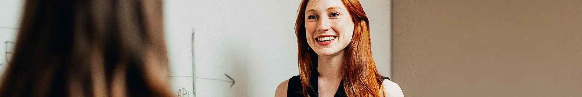 Das Foto zeigt eine Unterrichtssituation mit zwei Personen. Eine Person, deren Gesicht unscharf ist, blickt in die Kamera, während die andere von hinten zu sehen ist. Im Hintergrund befindet sich eine weiße Tafel mit einigen Schriftzeichen und einer einfachen Grafik mit einem nach rechts zeigenden Pfeil. Der Text auf der Tafel enthält „AP1“.