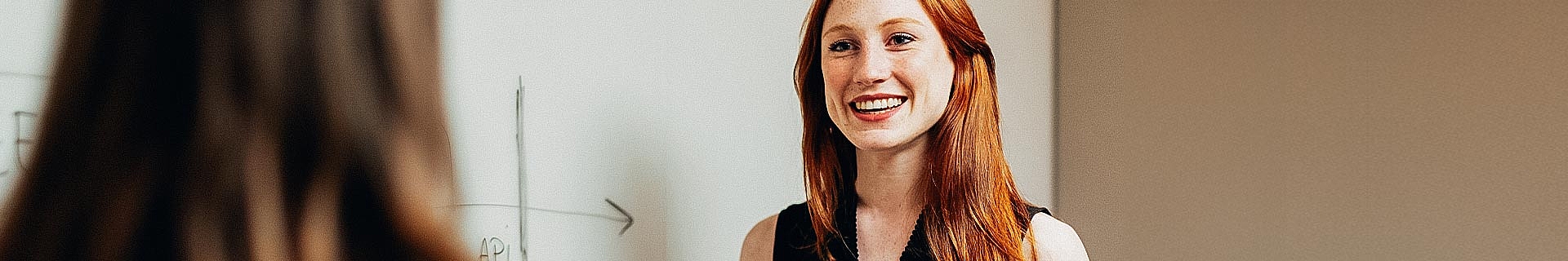 Das Foto zeigt eine Unterrichtssituation mit zwei Personen. Eine Person, deren Gesicht unscharf ist, blickt in die Kamera, während die andere von hinten zu sehen ist. Im Hintergrund befindet sich eine weiße Tafel mit einigen Schriftzeichen und einer einfachen Grafik mit einem nach rechts zeigenden Pfeil. Der Text auf der Tafel enthält „AP1“.