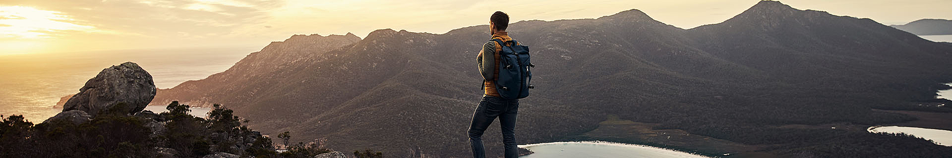 Das Bild zeigt eine Person, die auf einem felsigen Vorsprung steht und eine weite Landschaft aus Bergen und Tälern überblickt. Die Person trägt einen Rucksack und Outdoor-Kleidung, was auf eine Wanderung oder Erkundungstour hinweist. Die Szene spielt sich bei Sonnenaufgang oder Sonnenuntergang ab – der Himmel leuchtet in warmen Orangetönen und Gelbtönen.  Im Hintergrund erstrecken sich große Gebirgszüge, und in den Tälern ist etwas Vegetation zu erkennen. In der unteren rechten Ecke des Bildes ist teilweise ein Gewässer sichtbar.