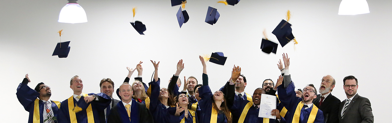 Das Bild zeigt Personen mit blauen Talaren und gelben Schärpen. wahrscheinlich Hochschulabsolventen. Sie werfen Akademikerhüte in die Luft. Einige Personen tragen Anzüge, wahrscheinlich Dozenten. 