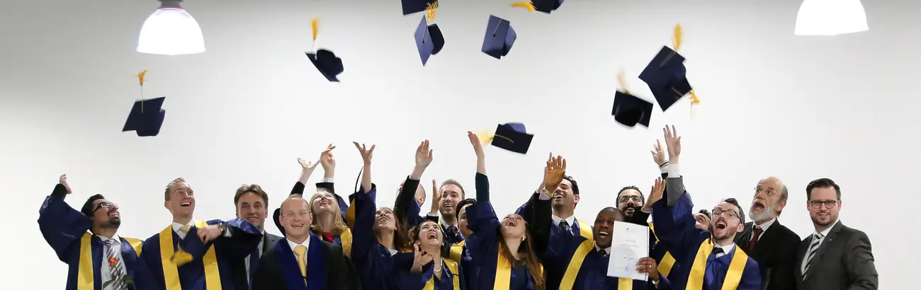 Das Bild zeigt Personen mit blauen Talaren und gelben Schärpen. wahrscheinlich Hochschulabsolventen. Sie werfen Akademikerhüte in die Luft. Einige Personen tragen Anzüge, wahrscheinlich Dozenten. 