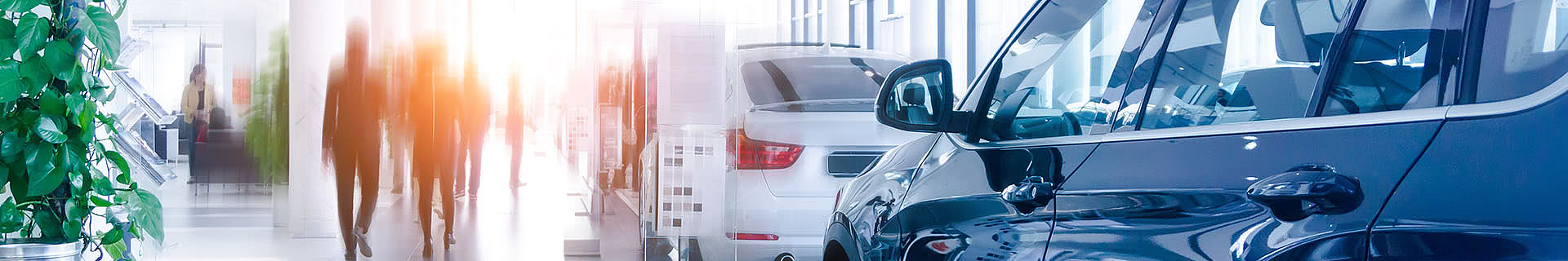 Das Bild zeigt das Innere eines modernen Autohauses. Auf der rechten Seite sind mehrere Fahrzeuge ausgestellt, darunter ein blaues Auto im Vordergrund und ein weißes weiter hinten. Auf der linken Seite befinden sich grüne Pflanzen sowie verschwommene Figuren von Personen, die sich bewegen oder stehen – ein Hinweis auf Aktivität im Autohaus. Im Hintergrund sorgen große Fenster für reichlich Tageslicht und eine helle, freundliche Atmosphäre.