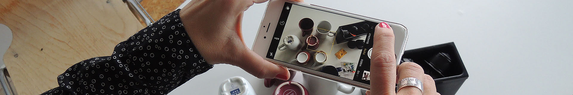 Das Bild zeigt eine Person, die mit einem Smartphone ein Foto von mehreren Gegenständen auf einem Tisch macht. Auf dem Tisch befinden sich verschiedene Tassen – einige mit Kaffee oder anderen Getränken – sowie weitere kleine Objekte. Die Hand der Person ist sichtbar, wobei ein Finger den Bildschirm des Smartphones berührt, um das Foto auszulösen. Die Person trägt ein schwarzes Shirt mit weißen Kreismustern und roten Nagellack auf den Nägeln. Im Hintergrund ist ein Teil eines Stuhls sowie eine offene schwarze Box auf dem Tisch zu sehen.