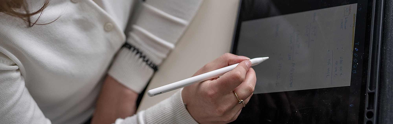 Das Bild zeigt eine Hand mit einem Stift vor einem Notizzettel. Es erinnert an eine Vorlesungssituation.