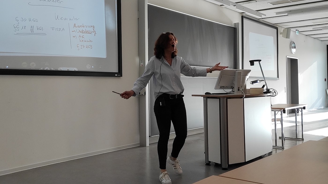 Professorin spricht gestikulierend vor Tafel.