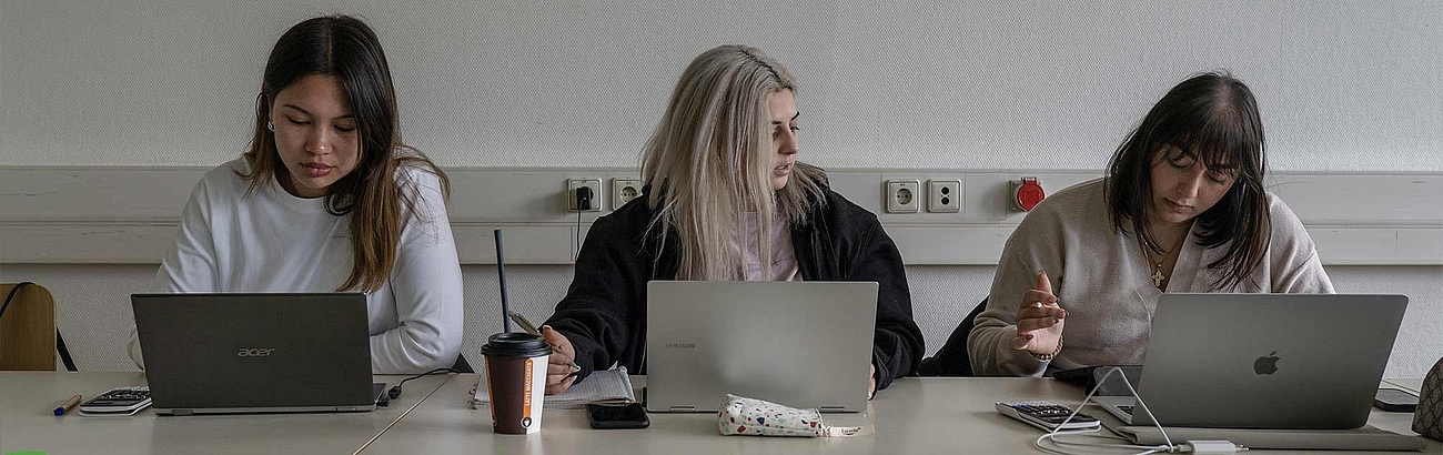 Das Bild zeigt drei Studentinnen an Laptops in einer Arbeitssituation.