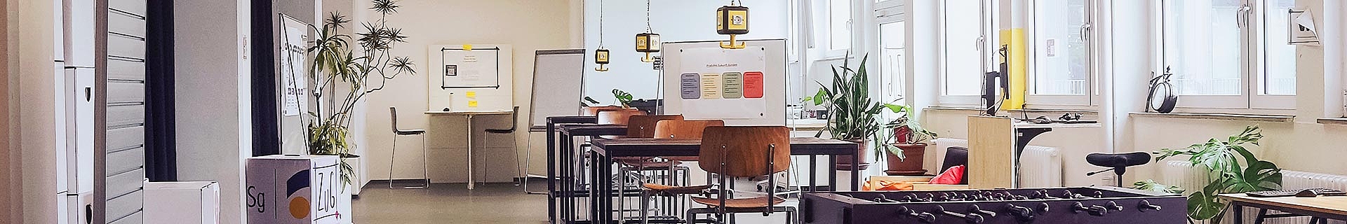 Das Bild zeigt einen modernen, gut beleuchteten Raum mit großen Fenstern auf der rechten Seite. In der Mitte stehen mehrere Tische und Stühle, darüber hängen Lampen. Auf der linken Seite befinden sich Spinde und eine große Pflanze. Im Hintergrund sieht man ein Whiteboard mit Text und Diagrammen sowie einen weiteren Tisch mit Stühlen. Im Vordergrund rechts ist ein Kickertisch zu erkennen.