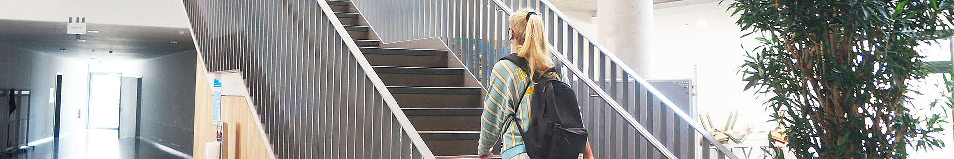 Das Bild zeigt eine Person mit langen blonden Haaren, die einen gestreiften Pullover trägt und einen schwarzen Rucksack auf dem Rücken hat. Sie geht eine Treppe hinauf in einem Innenraum. Die Treppe hat dunkle Stufen und Metallgeländer. Rechts neben der Treppe steht eine große Topfpflanze. Im Hintergrund ist ein Flur mit hellen Wänden und Böden zu sehen, der zu einem Bereich mit viel natürlichem Licht führt – vermutlich durch Fenster oder Türen am Ende des Korridors.