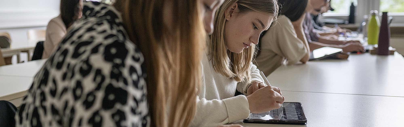 Das Bild zeigt zwei sitzende Studentinnen in einer Vorlesungssituation. Die Frauen machen sich Notizen. 