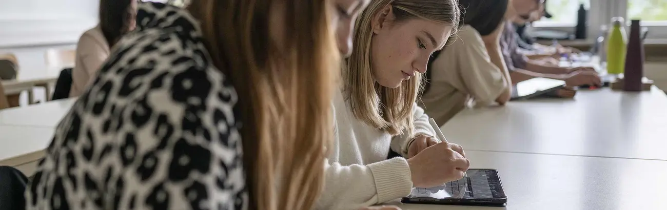 Das Bild zeigt zwei sitzende Studentinnen in einer Vorlesungssituation. Die Frauen machen sich Notizen. 