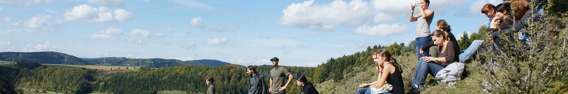 Das Bild zeigt eine Gruppe von Menschen, die auf einem grasbewachsenen Hügel sitzen und stehen. Im Hintergrund erstreckt sich eine malerische Landschaft mit sanften Hügeln und Wäldern. Der Himmel ist teilweise bewölkt, und die Szene vermittelt den Eindruck eines gemeinsamen Aufenthalts im Freien.
