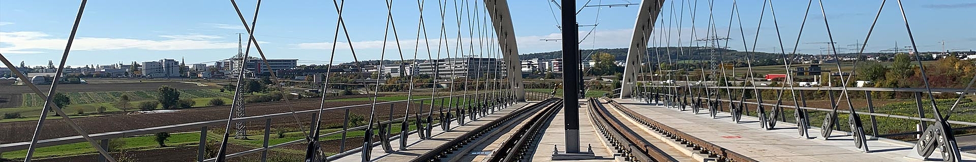 Das Bild zeigt eine Eisenbahnbrücke mit zwei parallel verlaufenden Gleisen. Die Brücke wird von zahlreichen vertikalen und diagonalen Kabeln getragen, die an einer Hauptstruktur aus großen Metallbögen befestigt sind. Im Hintergrund ist eine Landschaft mit Feldern, Bäumen und Gebäuden zu sehen, alles unter einem klaren blauen Himmel.