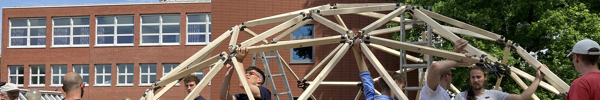Das Bild zeigt eine Gruppe von Personen, die eine geodätische Kuppel aus Holzträgern und Metallverbindern errichten. Im Hintergrund befindet sich ein rotes Backsteingebäude mit mehreren Fenstern. Auf der rechten Seite des Bildes sind Bäume zu sehen.