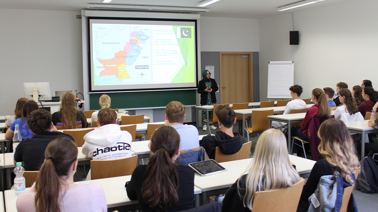 Schüler*innen hören einer internationalen Studentin zu, die in einem Klassenzimmer einen Vortrag hält.