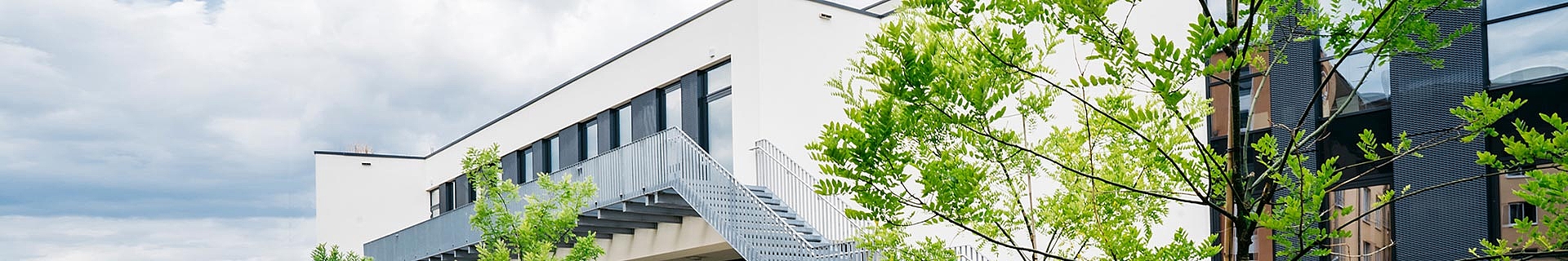 Das Bild zeigt ein modernes Gebäude mit weißer Fassade und großen Fenstern. Eine Außentreppe aus Metall führt zum oberen Stockwerk. Das Gebäude ist von grünen Bäumen umgeben, und der Himmel darüber ist bewölkt.