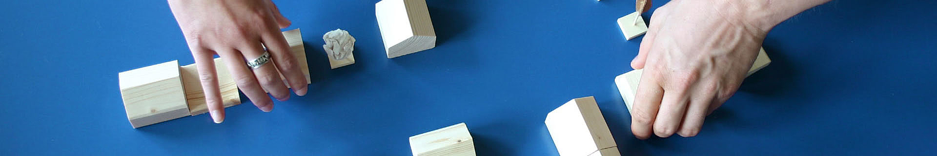 Das Bild zeigt zwei Hände, die mehrere kleine Holzklötze unterschiedlicher Formen und Größen auf einer blauen Oberfläche arrangieren oder bewegen. Zwischen den Klötzen liegt auch ein zerknülltes Stück Papier.  Das Bild steht symbolisch für kreatives Arbeiten, Problemlösung oder Planung.