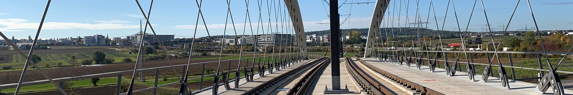 Das Bild zeigt eine Eisenbahnbrücke mit zwei parallel verlaufenden Gleisen. Die Brücke wird von zahlreichen vertikalen und diagonalen Kabeln getragen, die an einer Hauptstruktur aus großen Metallbögen befestigt sind. Im Hintergrund ist eine Landschaft mit Feldern, Bäumen und Gebäuden zu sehen, alles unter einem klaren blauen Himmel.