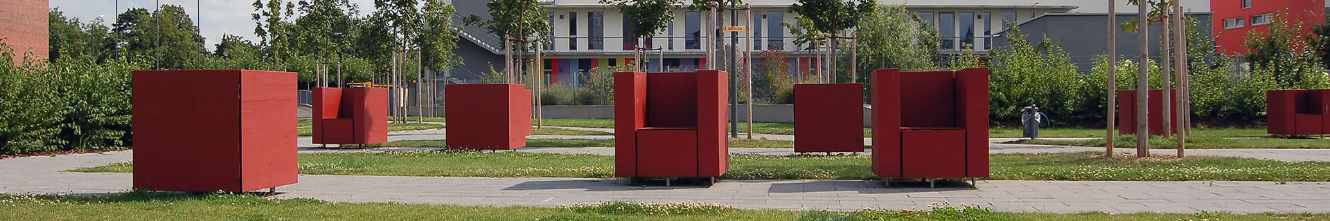 Das Bild zeigt einen Außenbereich mit mehreren großen, roten, würfelförmigen Strukturen, die wie Sitzgelegenheiten wirken. Diese sind auf einem gepflasterten Weg angeordnet, der von Rasenflächen und Bäumen umgeben ist. Im Hintergrund befindet sich ein modernes Gebäude mit vielen Fenstern und Balkonen. Die Szene wirkt wie ein Teil eines Parks oder öffentlichen Raums, der für Entspannung und soziale Begegnungen gestaltet wurde.