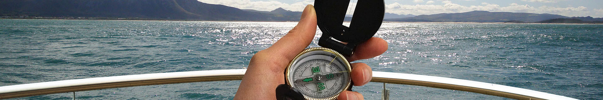 Das Bild zeigt eine Hand, die einen Kompass auf einem Boot hält. Im Hintergrund erstreckt sich das offene Meer, mit Bergen in der Ferne und einem teils bewölkten Himmel darüber. Der Kompass zeigt nach Norden, und im Vordergrund ist das Geländer des Boots zu sehen. Das Bild vermittelt ein Gefühl von Orientierung, Navigation und Abenteuer. 